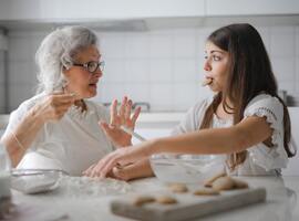 Eine ältere Frau, links im Bild, sitzt mit ihrer Enkelin, rechts im Bild, in der Küche, umgeben von Backutensilien und frisch gebackenen Keksen.
