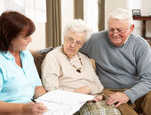 Ein Seniorenpaar sitzt gemeinsam mit einer Altenpflegerin auf der heimischen Couch. Die Altenpflegerin erklärt etwas anhand von Notizen in einem Ordner.