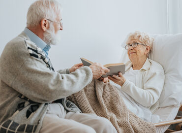 Senior reicht seiner Partnerin, die in einem Pflegebett liegt, ein aufgeschlagenes Buch. Beide lächeln.